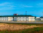 Medium distant photograph of the Virginia Center for Behavioral Rehabilitation (VCBR) in Burkeville, Virginia. This is Virginia's Shadow Prison. The image shows brown grass in the foreground, with an institutional prison-like building behinds a tall double razor wire fence that wraps over into the compound for the shadow prison, similar to the fence use to contain velociraptors in the original Jurassic Park film.